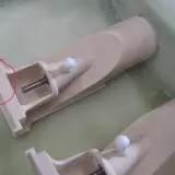 Zwei Teile eines wellenantrieb d-drive Systems, beige, mit Metallachsen und weißen Kugeln, liegen in einer grünen Flüssigkeit.