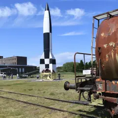 Eine V2-Rakete, ein Symbol deutscher Raketentechnik im 2. Weltkrieg, steht auf einem Freigelände neben einem rostigen Waggon.