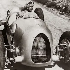 Ein Rennfahrer in einem stromlinienförmigen Auto, ein deutscher Kfz-Pionier, fährt auf einer Schotterstraße.