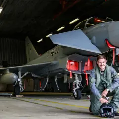 Ein junger F16 Pilot in Uniform kniet neben seinem Jet in einer Flugzeughalle.