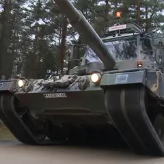 Ein Leopard 1 Panzer fährt durch einen Wald. Auf dem Dach ist ein Schild mit "FAHRSCHULE" angebracht.
