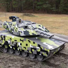 Ein moderner Jagdpanzer mit digitaler Tarnung steht auf schlammigem Untergrund vor einem Wald.