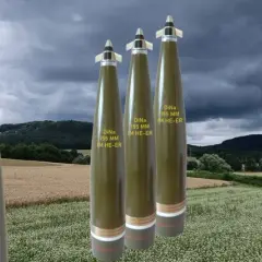 Vier Geschosse des Kalibers 155 mm stehen im Vordergrund vor einer ländlichen Kulisse mit Feldern und bewaldeten Hügeln unter dramatischem Himmel.
