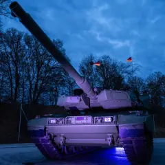 Ein moderner Leopard 2 panzer steht im Dunkeln, beleuchtet von blauem Licht. Deutsche Flaggen wehen im Hintergrund.