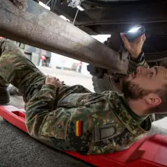 Ein Soldat, ein Unteroffizier ohne Portepee, prüft mit einer Lampe das Fahrgestell eines Fahrzeugs.