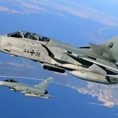 Zwei deutsche Kampfflugzeuge, ein PA-200 Tornado und ein Eurofighter, fliegen über Küstenlandschaft.