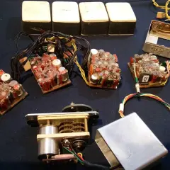 Elektronikbauteile, darunter vier weiße Kästen, mehrere Leiterplatten mit vielen Kondensatoren und Drähten, ein Motor mit Zahnrädern und ein Metallgehäuse. Vielleicht für 27 MHz Funkgeräte.