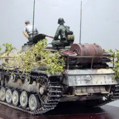 Sturmhaubitze 42 Modell mit zwei Figuren, getarnt mit Zweigen.