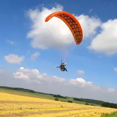 Ein Gleitschirmpilot mit einem orangen hepf micro vario Schirm fliegt über ein gelbes Feld unter blauem Himmel mit Wolken.