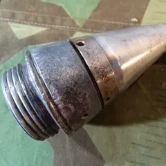 Ein metallisches Teil, das an eine **Flakgranate** erinnert, mit Gewinde und spitz zulaufendem Ende, liegt auf Tarnstoff.