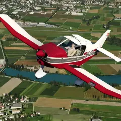 Ein roter und weißer Robin DR 400 fliegt über grüne Felder und einen Fluss.