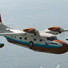 Grumman Mallard Wasserflugzeug fliegt über bewaldetem Gebiet. Das Flugzeug hat eine weiße und braune Lackierung mit blauen Akzenten.