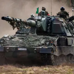 Eine Panzerhaubitze 3000 mit Tarnnetz fährt durch staubiges Gelände. Zwei Soldaten sind auf dem Turm zu sehen.