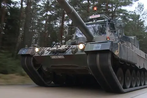 Ein Leopard 1 Panzer fährt durch einen Wald. Auf dem Dach ist ein Schild mit "FAHRSCHULE" angebracht.