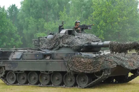 Ein Leopard 1 Panzer mit Tarnnetz und einem Soldaten am Maschinengewehr. Sein Gewicht ist beeindruckend.