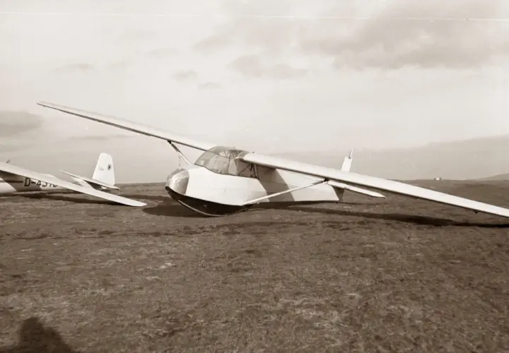 Ein Segelflugzeug vom Typ Musger MG 19 steht auf einer Wiese. Ein weiteres Flugzeug ist im Hintergrund zu sehen.