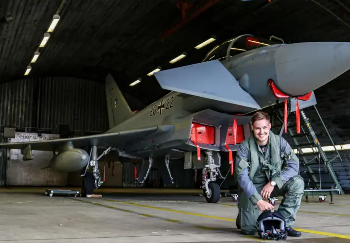 Ein junger F16 Pilot in Uniform kniet neben seinem Jet in einer Flugzeughalle.