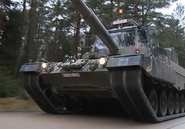 Ein Leopard 1 Panzer fährt durch einen Wald. Auf dem Dach ist ein Schild mit "FAHRSCHULE" angebracht.