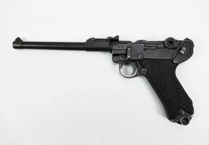 Eine schwarze Luger Pistole mit nummeriertem Schlitten (938) und geriffeltem Griff liegt auf weißem Grund.