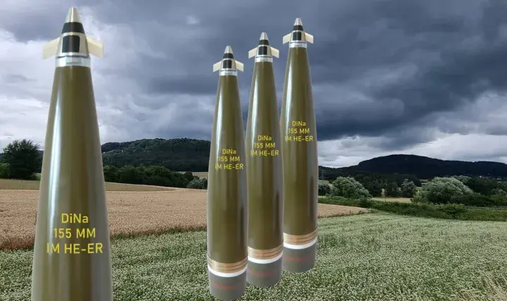 Vier Geschosse des Kalibers 155 mm stehen im Vordergrund vor einer ländlichen Kulisse mit Feldern und bewaldeten Hügeln unter dramatischem Himmel.