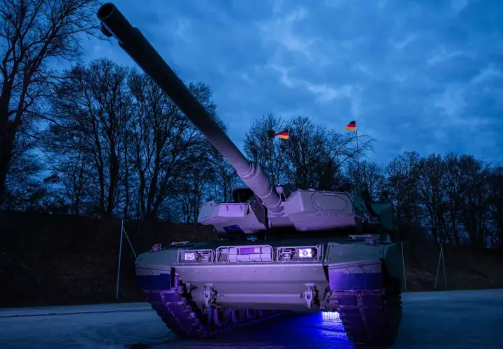 Ein moderner Leopard 2 panzer steht im Dunkeln, beleuchtet von blauem Licht. Deutsche Flaggen wehen im Hintergrund.
