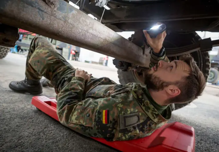 Ein Soldat, ein Unteroffizier ohne Portepee, prüft mit einer Lampe das Fahrgestell eines Fahrzeugs.