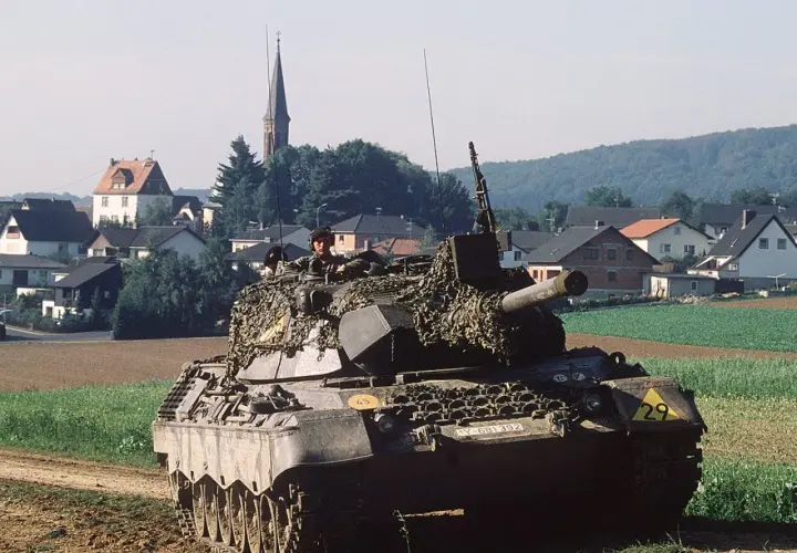 Ein Leopard 1 Panzer mit Tarnnetz fährt durch ein Feld. Die Munition ist bereit für den Einsatz.
