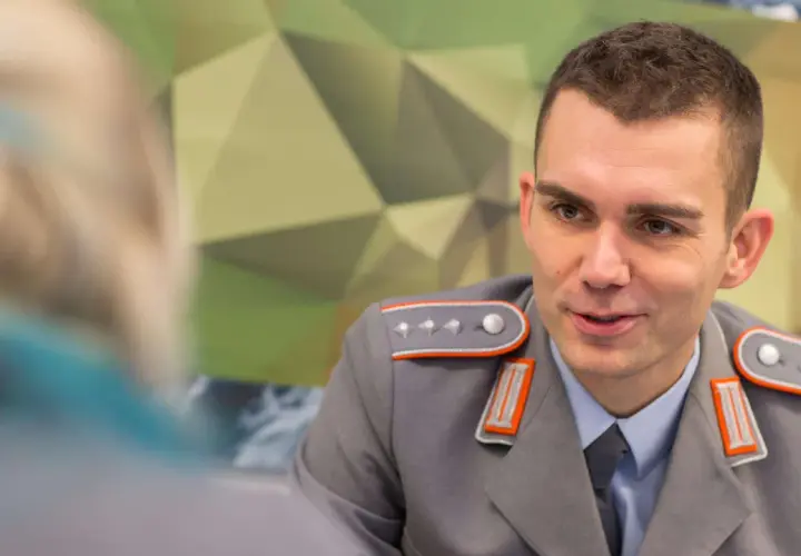 Ein junger Fachoffizier der Bundeswehr im Gespräch, trägt eine graue Uniform mit orangefarbenen Akzenten und Schulterklappen.