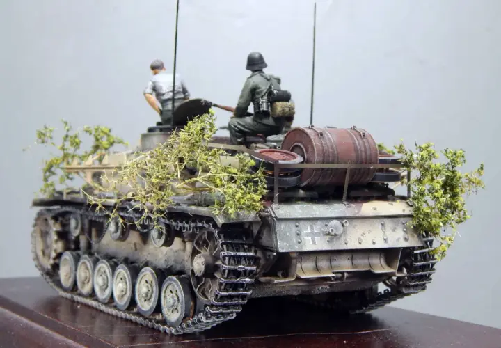 Sturmhaubitze 42 Modell mit zwei Figuren, getarnt mit Zweigen.