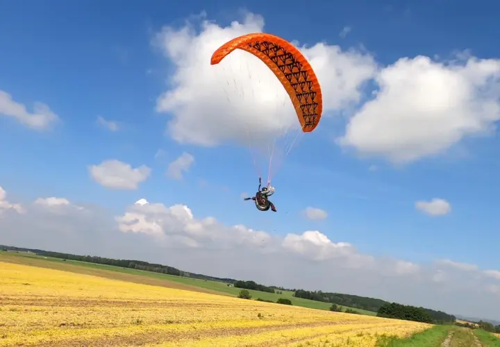 Ein Gleitschirmpilot mit einem orangen hepf micro vario Schirm fliegt über ein gelbes Feld unter blauem Himmel mit Wolken.