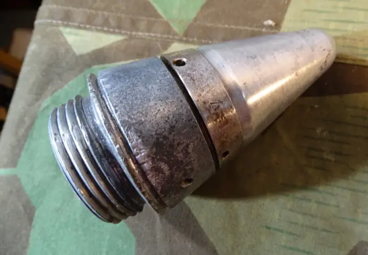 Ein metallisches Teil, das an eine **Flakgranate** erinnert, mit Gewinde und spitz zulaufendem Ende, liegt auf Tarnstoff.