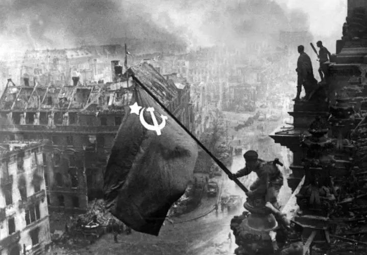 Sowjetische Soldaten hissen die rote Fahne über dem zerstörten Reichstag 1945. Rauch und Trümmer prägen das Bild Berlins.
