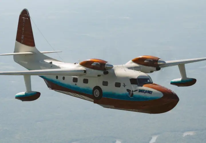 Grumman Mallard Wasserflugzeug fliegt über bewaldetem Gebiet. Das Flugzeug hat eine weiße und braune Lackierung mit blauen Akzenten.