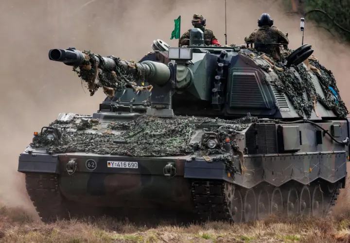 Eine Panzerhaubitze 3000 mit Tarnnetz fährt durch staubiges Gelände. Zwei Soldaten sind auf dem Turm zu sehen.