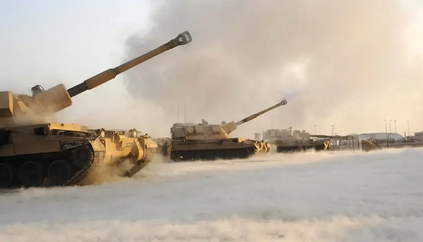 Mehrere Panzerhaubitzen, wie die 90, feuern in einer staubigen, rauchigen Umgebung.