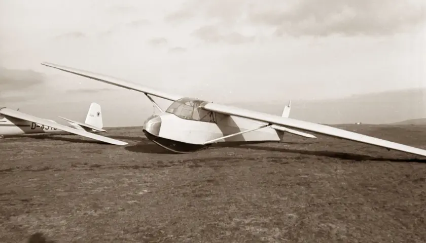 Ein Segelflugzeug vom Typ Musger MG 19 steht auf einer Wiese. Ein weiteres Flugzeug ist im Hintergrund zu sehen.