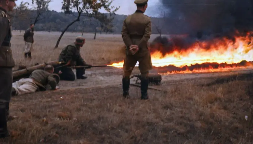 Soldaten im 2. Weltkrieg üben den Einsatz eines Flammenwerfers. Ein Mann zielt mit dem Gerät, während ein anderer auf dem Boden liegt.