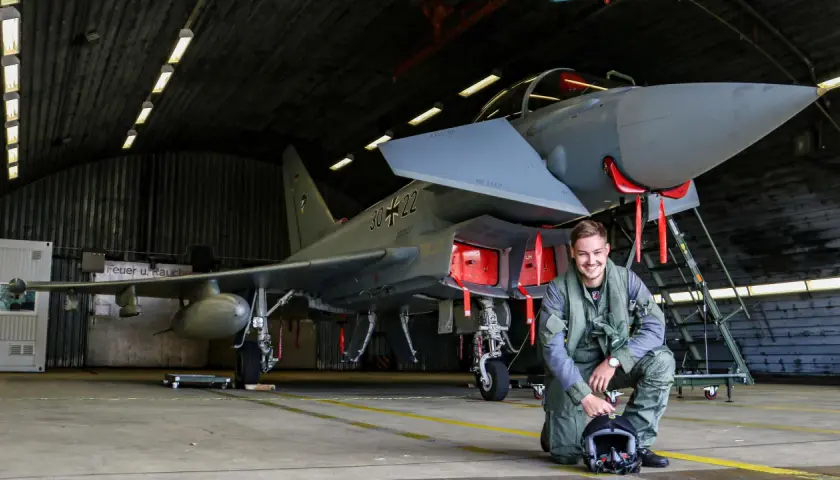 Ein junger F16 Pilot in Uniform kniet neben seinem Jet in einer Flugzeughalle.