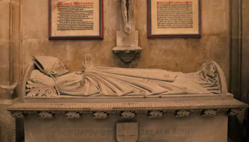 Das Michael Wittmann Grab zeigt eine liegende Steinfigur eines Bischofs mit gefalteten Händen. Tafeln mit Texten flankieren die Skulptur.