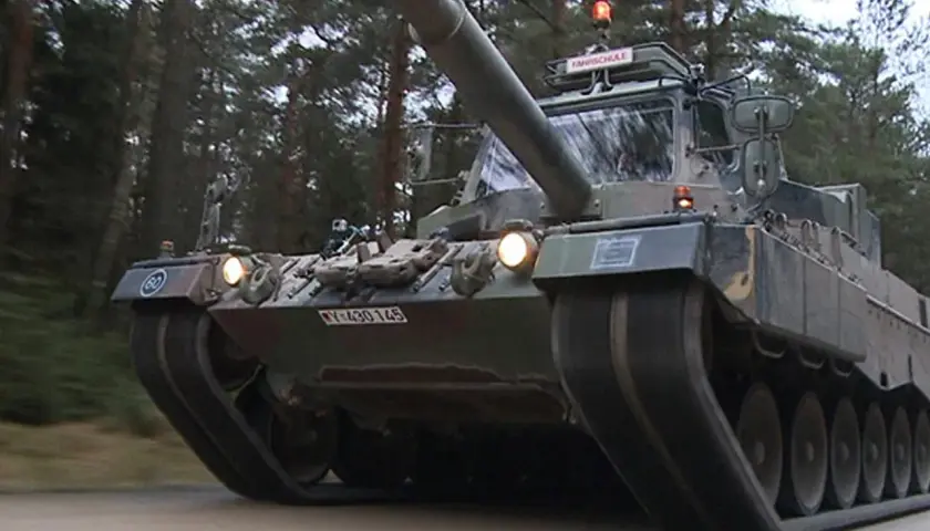 Ein Leopard 1 Panzer fährt durch einen Wald. Auf dem Dach ist ein Schild mit "FAHRSCHULE" angebracht.