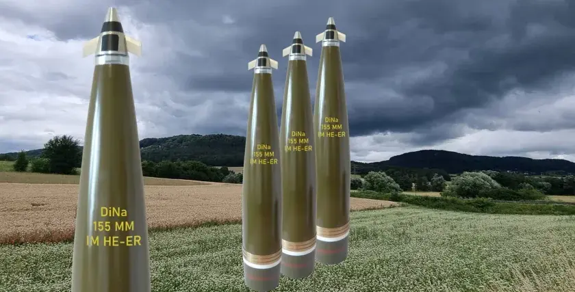 Vier Geschosse des Kalibers 155 mm stehen im Vordergrund vor einer ländlichen Kulisse mit Feldern und bewaldeten Hügeln unter dramatischem Himmel.