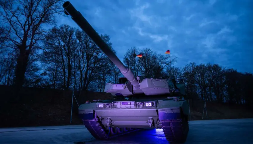 Ein moderner Leopard 2 panzer steht im Dunkeln, beleuchtet von blauem Licht. Deutsche Flaggen wehen im Hintergrund.