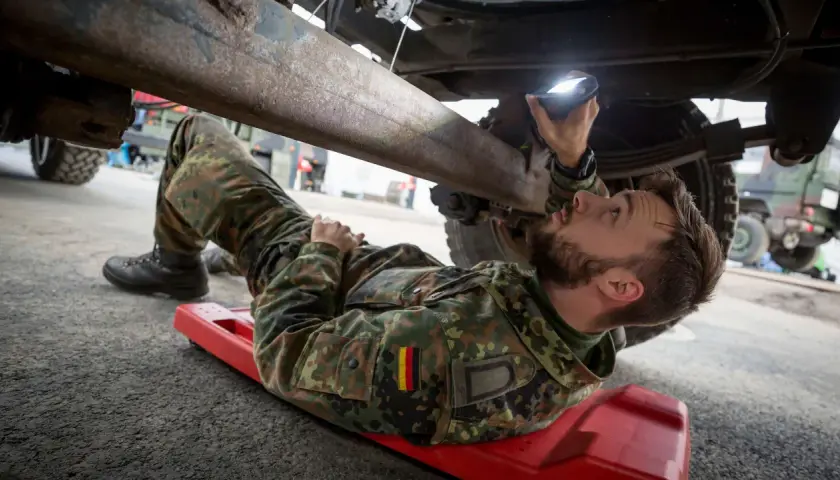 Ein Soldat, ein Unteroffizier ohne Portepee, prüft mit einer Lampe das Fahrgestell eines Fahrzeugs.