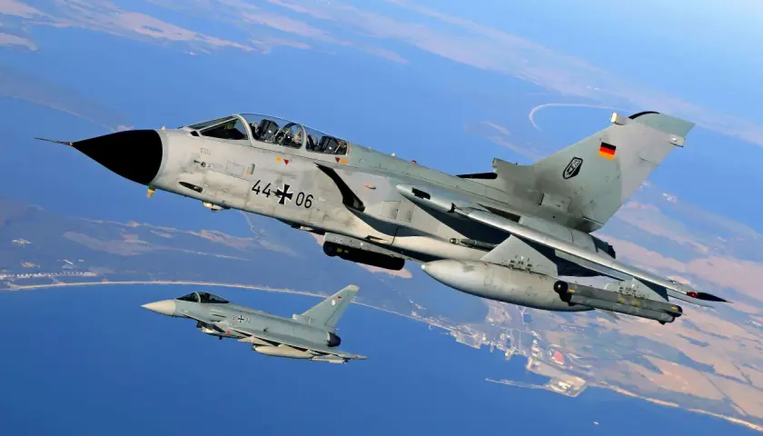 Zwei deutsche Kampfflugzeuge, ein PA-200 Tornado und ein Eurofighter, fliegen über Küstenlandschaft.