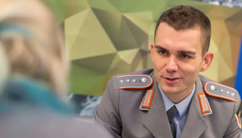 Ein junger Fachoffizier der Bundeswehr im Gespräch, trägt eine graue Uniform mit orangefarbenen Akzenten und Schulterklappen.
