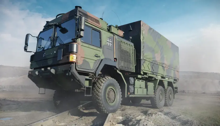 Ein gepanzerter LKW der Bundeswehr mit Tarnmuster fährt über Schienen in einer staubigen Landschaft.
