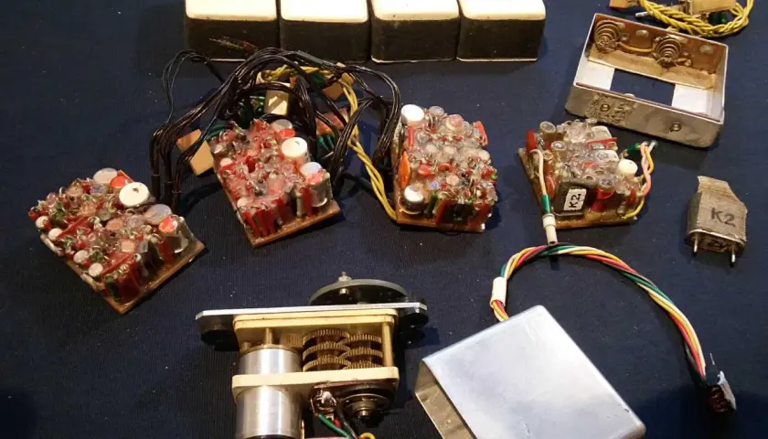Elektronikbauteile, darunter vier weiße Kästen, mehrere Leiterplatten mit vielen Kondensatoren und Drähten, ein Motor mit Zahnrädern und ein Metallgehäuse. Vielleicht für 27 MHz Funkgeräte.