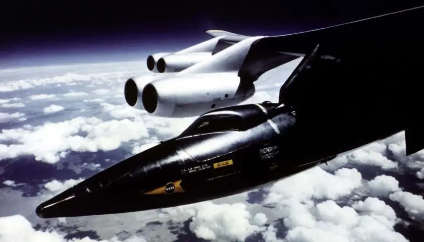 Das X-15 Flugzeug, ein schwarzer Pfeil, fliegt über Wolken. Ein größeres, weißes Flugzeug mit zwei Triebwerken ist daneben zu sehen.