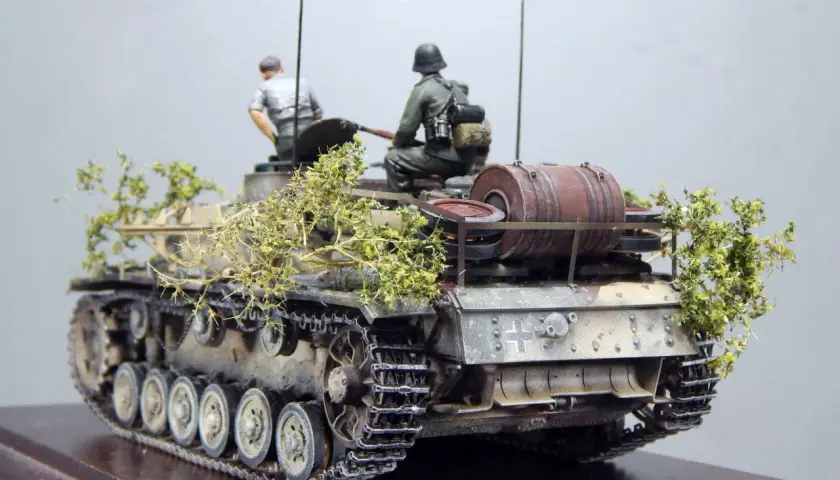 Sturmhaubitze 42 Modell mit zwei Figuren, getarnt mit Zweigen.