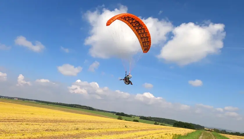 Ein Gleitschirmpilot mit einem orangen hepf micro vario Schirm fliegt über ein gelbes Feld unter blauem Himmel mit Wolken.